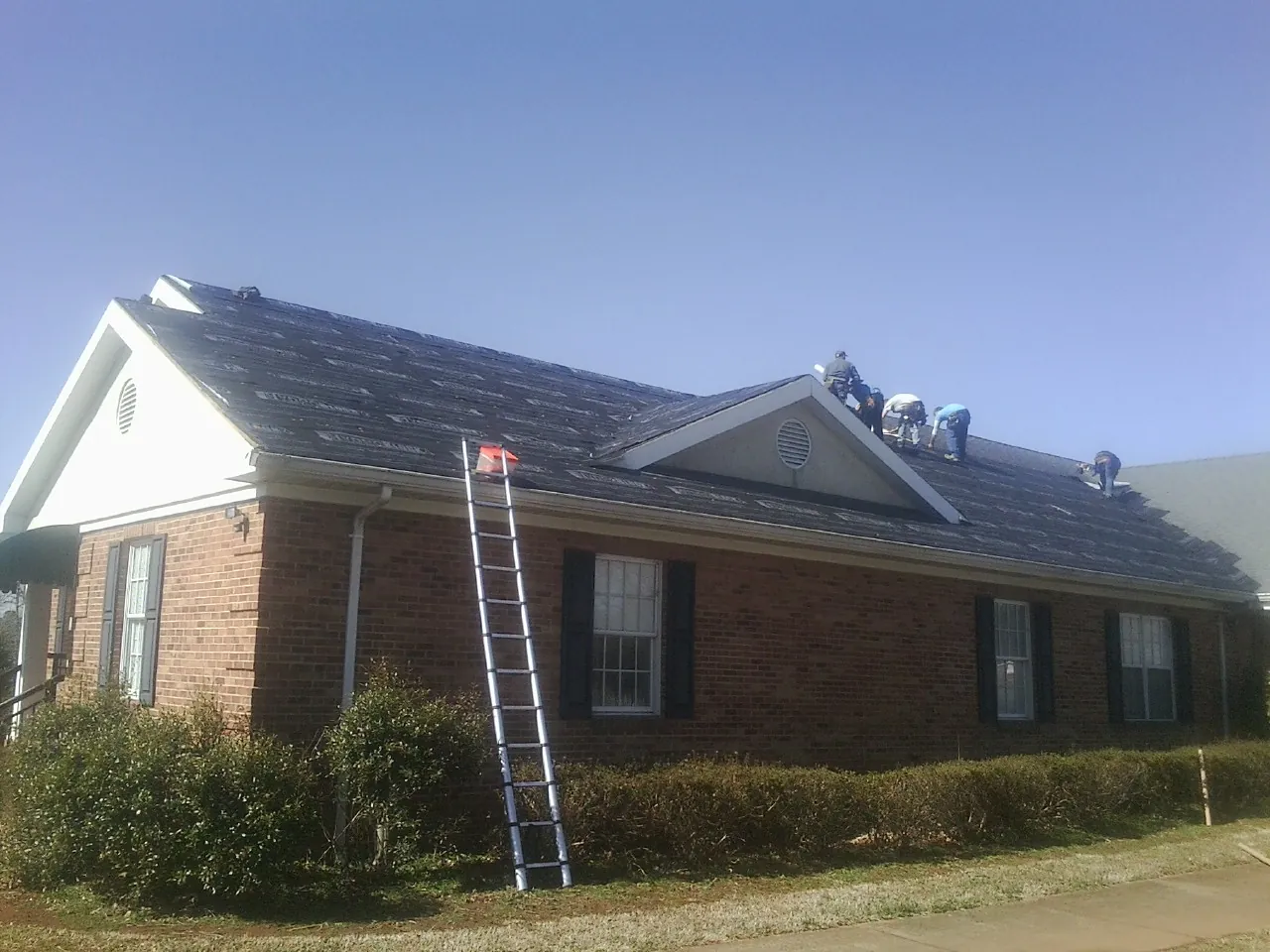 Crew tearing shingles off a brick ranch roof with a ladder set — Emergency Roof Repair work by our crew in Ferris
