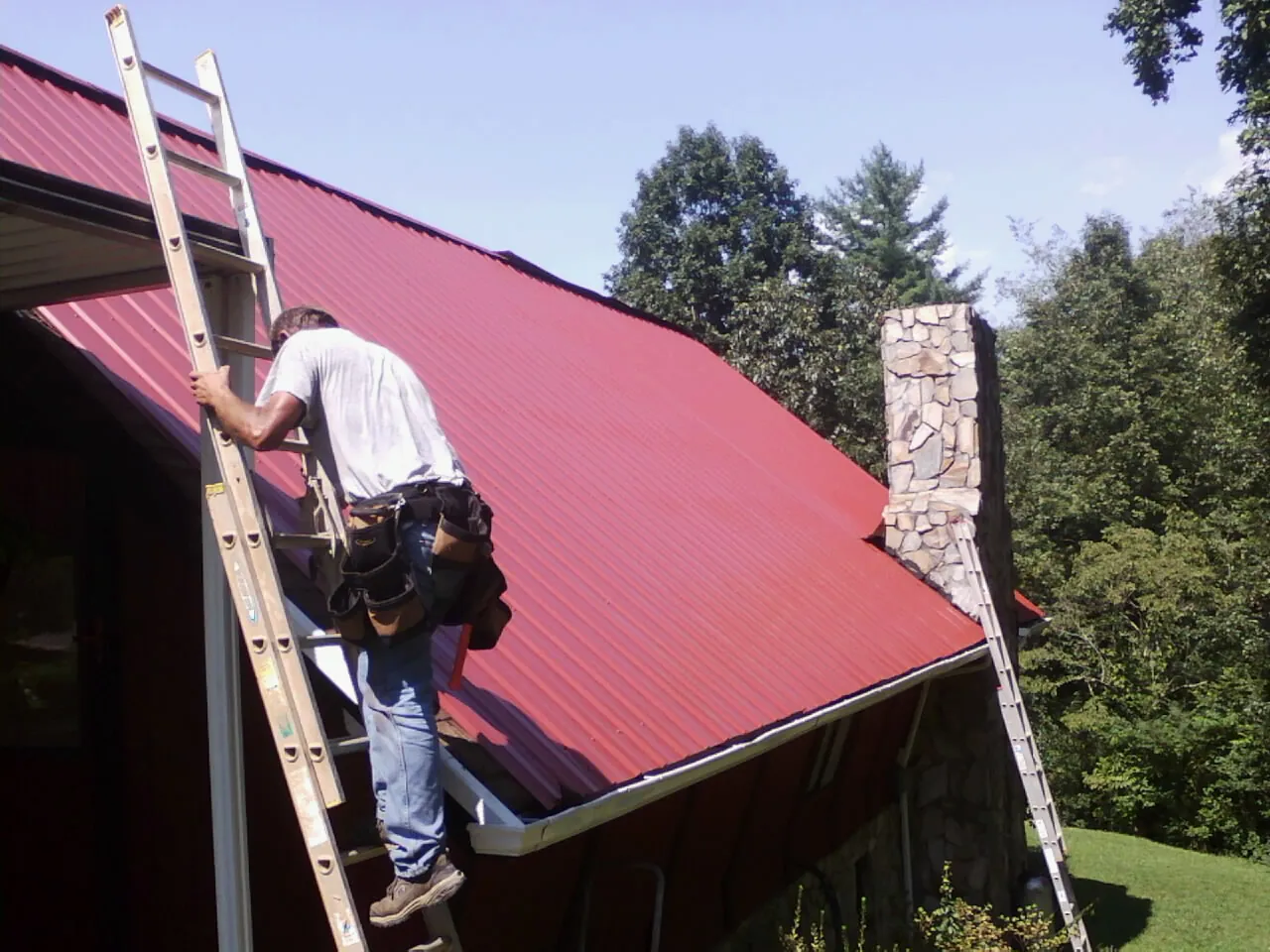 Roofer on a ladder beside a red metal roof and stone chimney — Soffit & Fascia Repair by Roofing Brothers in Ferris